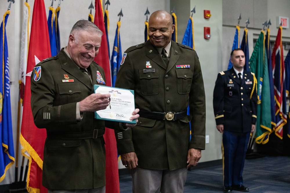 Two senior U.S. Army officers in olive green dress uniforms are participating in a military awards ceremony, with one officer holding a certificate or commendation document. Both men are smiling, suggesting a positive and celebratory occasion. A third soldier stands at attention in the background amid a display of multiple military unit and organizational flags.