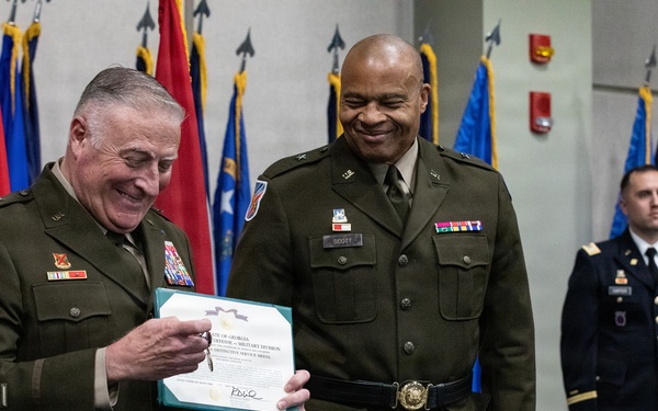 Two senior U.S. Army officers in olive green dress uniforms are participating in a military awards ceremony, with one officer holding a certificate or commendation document. Both men are smiling, suggesting a positive and celebratory occasion. A third soldier stands at attention in the background amid a display of multiple military unit and organizational flags.