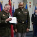 Two senior U.S. Army officers in olive green dress uniforms are participating in a military awards ceremony, with one officer holding a certificate or commendation document. Both men are smiling, suggesting a positive and celebratory occasion. A third soldier stands at attention in the background amid a display of multiple military unit and organizational flags.