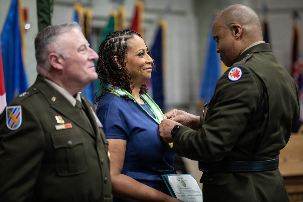 A military ceremony is taking place in which a uniformed officer is presenting or pinning a medal/award to a civilian woman wearing a green and white ribbon around her neck. A second uniformed officer stands to the left observing the ceremony with a smile. Multiple national and organizational flags are visible in the background.