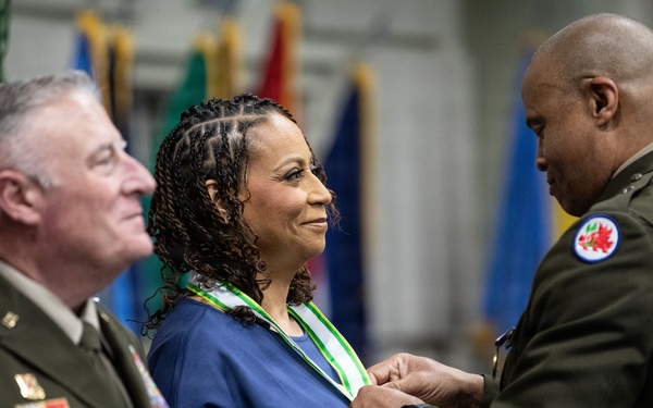 A military ceremony is taking place in which a uniformed officer is presenting or pinning a medal/award to a civilian woman wearing a green and white ribbon around her neck. A second uniformed officer stands to the left observing the ceremony with a smile. Multiple national and organizational flags are visible in the background.