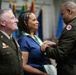 A military ceremony is taking place in which a uniformed officer is presenting or pinning a medal/award to a civilian woman wearing a green and white ribbon around her neck. A second uniformed officer stands to the left observing the ceremony with a smile. Multiple national and organizational flags are visible in the background.