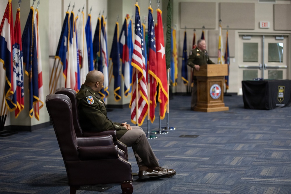 A formal U.S. Army military ceremony is taking place in an official hall lined with numerous state and military flags. One officer in Army Service Uniform sits in a leather armchair in the foreground, while another officer speaks at a wooden podium in the background. The setting and arrangement suggest a change of command, promotion, or retirement ceremony.