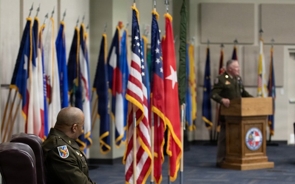 A formal U.S. Army military ceremony is taking place in an official hall lined with numerous state and military flags. One officer in Army Service Uniform sits in a leather armchair in the foreground, while another officer speaks at a wooden podium in the background. The setting and arrangement suggest a change of command, promotion, or retirement ceremony.