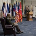 A formal U.S. Army military ceremony is taking place in an official hall lined with numerous state and military flags. One officer in Army Service Uniform sits in a leather armchair in the foreground, while another officer speaks at a wooden podium in the background. The setting and arrangement suggest a change of command, promotion, or retirement ceremony.