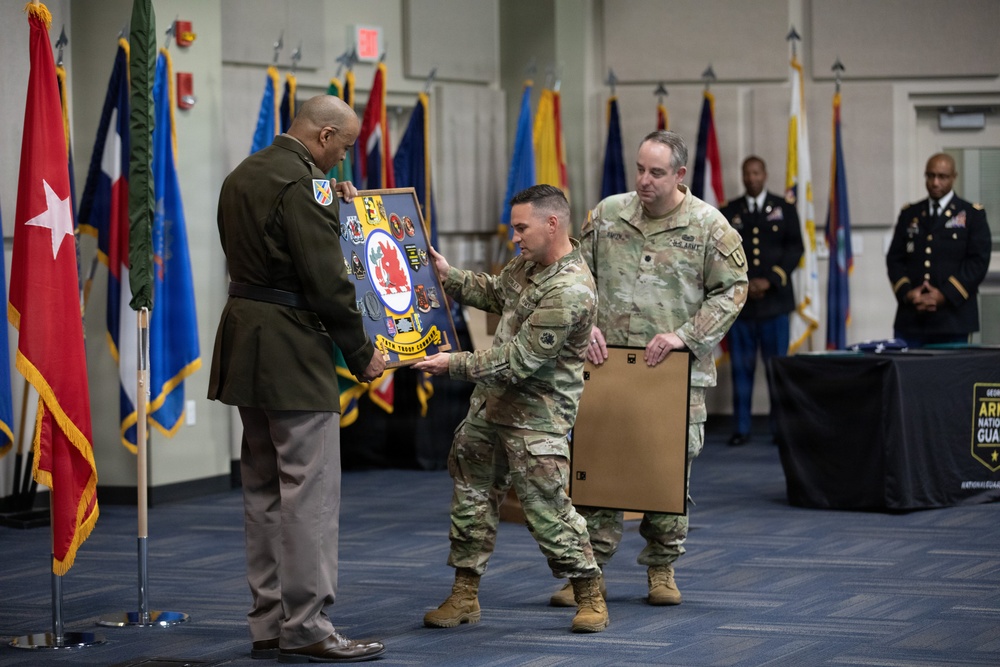 A military ceremony is taking place in a formal indoor venue, likely a change of command or retirement ceremony. Two soldiers in Army Combat Uniforms (ACU/OCP pattern) are presenting a shadow box or unit crest display board to a senior officer in dress uniform. The setting includes multiple military flags and a Georgia Army National Guard banner visible in the background.