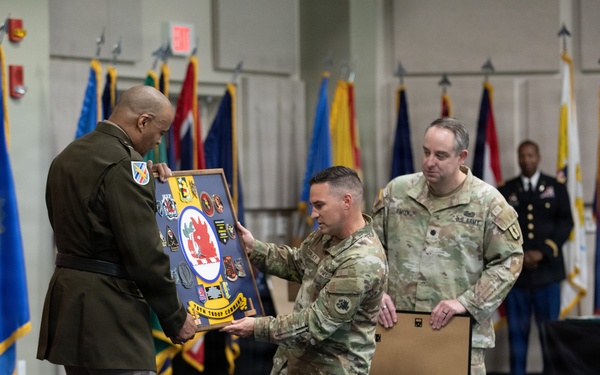 A military ceremony is taking place in a formal indoor venue, likely a change of command or retirement ceremony. Two soldiers in Army Combat Uniforms (ACU/OCP pattern) are presenting a shadow box or unit crest display board to a senior officer in dress uniform. The setting includes multiple military flags and a Georgia Army National Guard banner visible in the background.
