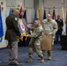 A military ceremony is taking place in a formal indoor venue, likely a change of command or retirement ceremony. Two soldiers in Army Combat Uniforms (ACU/OCP pattern) are presenting a shadow box or unit crest display board to a senior officer in dress uniform. The setting includes multiple military flags and a Georgia Army National Guard banner visible in the background.