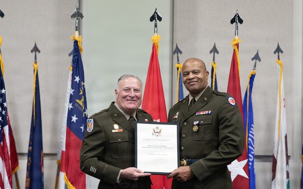 Two U.S. Army officers in olive green service uniforms are posing together while jointly holding an official certificate or commendation document. The ceremony appears to be a formal military award or recognition presentation, taking place in front of an array of U.S. state and military flags. Both individuals are smiling, suggesting a positive ceremonial occasion.