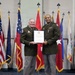 Two U.S. Army officers in olive green service uniforms are posing together while jointly holding an official certificate or commendation document. The ceremony appears to be a formal military award or recognition presentation, taking place in front of an array of U.S. state and military flags. Both individuals are smiling, suggesting a positive ceremonial occasion.