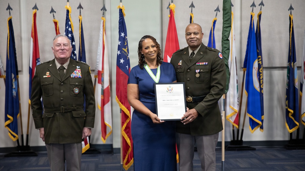 Three individuals pose for a formal military ceremony photograph in front of a display of multiple flags. A woman in a blue dress wearing a medallion/ribbon award holds a framed certificate, flanked by two military officers in Army dress uniforms. The setting appears to be an official military recognition or award ceremony.