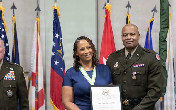 Three individuals pose for a formal military ceremony photograph in front of a display of multiple flags. A woman in a blue dress wearing a medallion/ribbon award holds a framed certificate, flanked by two military officers in Army dress uniforms. The setting appears to be an official military recognition or award ceremony.