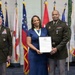 Three individuals pose for a formal military ceremony photograph in front of a display of multiple flags. A woman in a blue dress wearing a medallion/ribbon award holds a framed certificate, flanked by two military officers in Army dress uniforms. The setting appears to be an official military recognition or award ceremony.