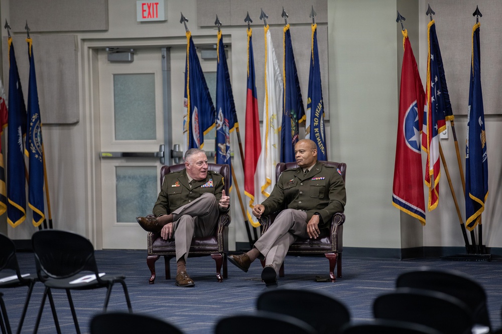 Two military officers in olive green dress uniforms are seated in leather armchairs on a stage or presentation area, engaged in conversation. They are positioned in front of a display of multiple state and organizational flags. The setting appears to be a formal military event or briefing room with rows of empty chairs in the foreground.