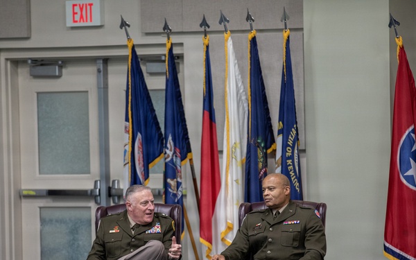 Two military officers in olive green dress uniforms are seated in leather armchairs on a stage or presentation area, engaged in conversation. They are positioned in front of a display of multiple state and organizational flags. The setting appears to be a formal military event or briefing room with rows of empty chairs in the foreground.