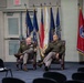 Two military officers in olive green dress uniforms are seated in leather armchairs on a stage or presentation area, engaged in conversation. They are positioned in front of a display of multiple state and organizational flags. The setting appears to be a formal military event or briefing room with rows of empty chairs in the foreground.