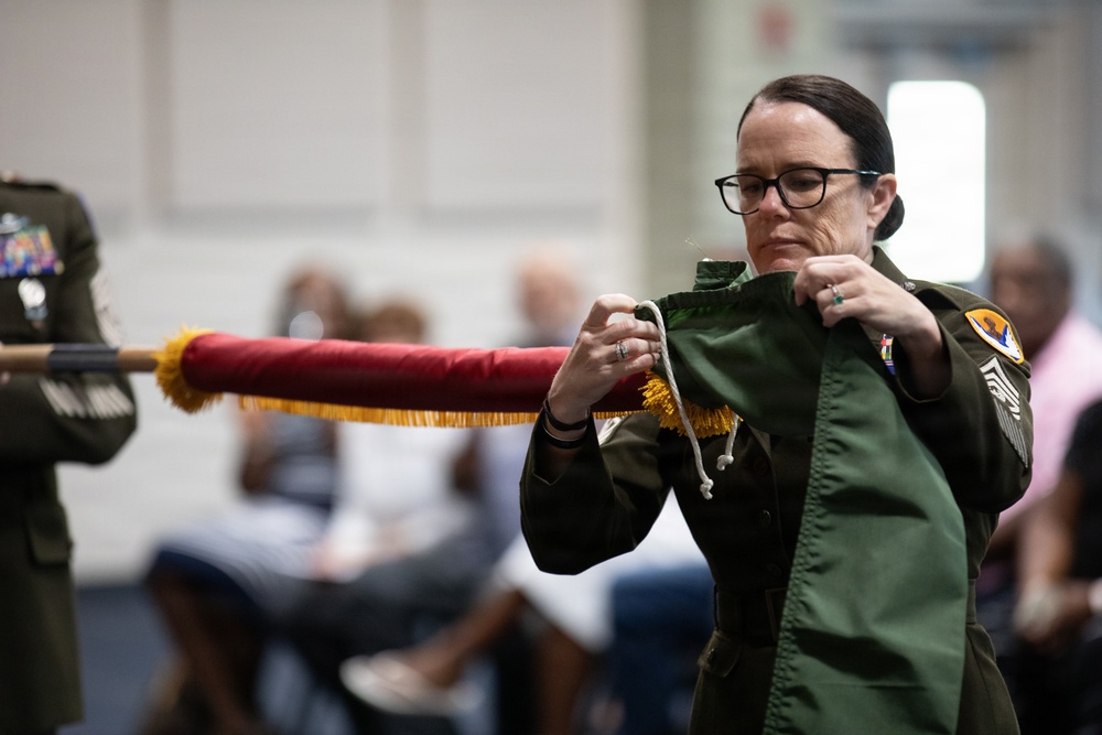 A female military service member in a green flight suit is handling a furled guidon or ceremonial flag with red fabric and gold fringe, appearing to tie or untie cords securing it. She wears glasses, has her hair pulled back, and displays visible rank insignia and unit patches on her uniform. The setting appears to be an indoor military ceremony with seated audience members and another uniformed individual visible in the background.