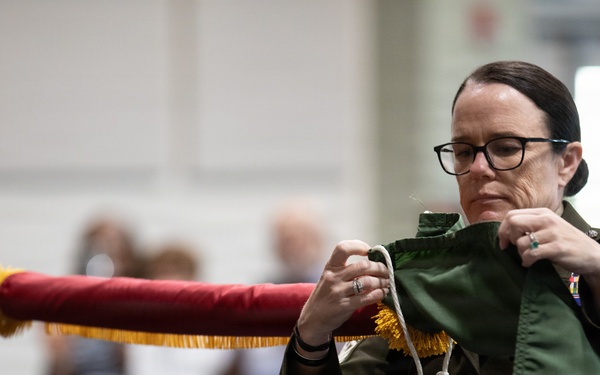 A female military service member in a green flight suit is handling a furled guidon or ceremonial flag with red fabric and gold fringe, appearing to tie or untie cords securing it. She wears glasses, has her hair pulled back, and displays visible rank insignia and unit patches on her uniform. The setting appears to be an indoor military ceremony with seated audience members and another uniformed individual visible in the background.