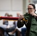A female military service member in a green flight suit is handling a furled guidon or ceremonial flag with red fabric and gold fringe, appearing to tie or untie cords securing it. She wears glasses, has her hair pulled back, and displays visible rank insignia and unit patches on her uniform. The setting appears to be an indoor military ceremony with seated audience members and another uniformed individual visible in the background.