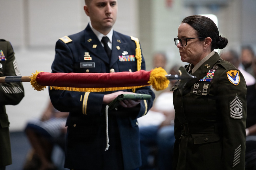 A military ceremony is taking place involving at least two uniformed service members. A male soldier wearing a blue Army Service Uniform with a name tag reading 'CARTER' and a female soldier in an olive green Army uniform with Command Sergeant Major rank insignia are handling a furled guidon or organizational color with red fabric and gold fringe. The setting appears to be an indoor venue with seated civilians in the background.