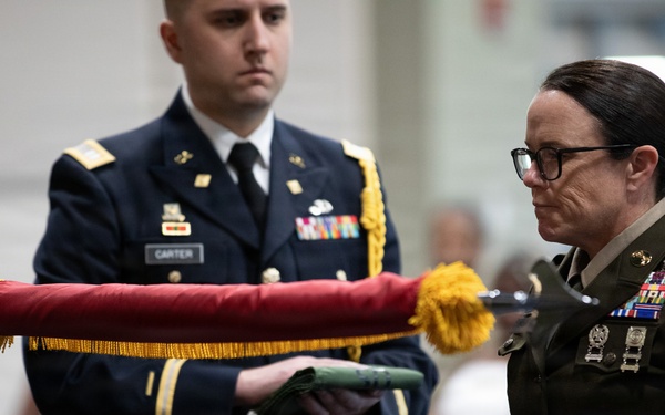 A military ceremony is taking place involving at least two uniformed service members. A male soldier wearing a blue Army Service Uniform with a name tag reading 'CARTER' and a female soldier in an olive green Army uniform with Command Sergeant Major rank insignia are handling a furled guidon or organizational color with red fabric and gold fringe. The setting appears to be an indoor venue with seated civilians in the background.