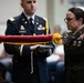 A military ceremony is taking place involving at least two uniformed service members. A male soldier wearing a blue Army Service Uniform with a name tag reading 'CARTER' and a female soldier in an olive green Army uniform with Command Sergeant Major rank insignia are handling a furled guidon or organizational color with red fabric and gold fringe. The setting appears to be an indoor venue with seated civilians in the background.