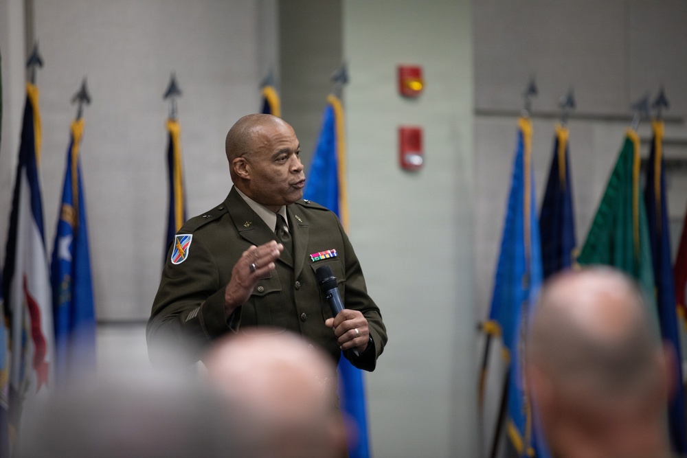 A U.S. Army officer in dress green uniform is speaking at a formal military event, holding a microphone and addressing an audience. Multiple military flags are displayed in the background, suggesting an official ceremony or briefing. The officer wears visible rank insignia, a unit patch, and service ribbons on his uniform.