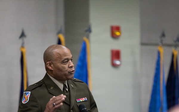 A U.S. Army officer in dress green uniform is speaking at a formal military event, holding a microphone and addressing an audience. Multiple military flags are displayed in the background, suggesting an official ceremony or briefing. The officer wears visible rank insignia, a unit patch, and service ribbons on his uniform.
