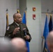 A U.S. Army officer in dress green uniform is speaking at a formal military event, holding a microphone and addressing an audience. Multiple military flags are displayed in the background, suggesting an official ceremony or briefing. The officer wears visible rank insignia, a unit patch, and service ribbons on his uniform.