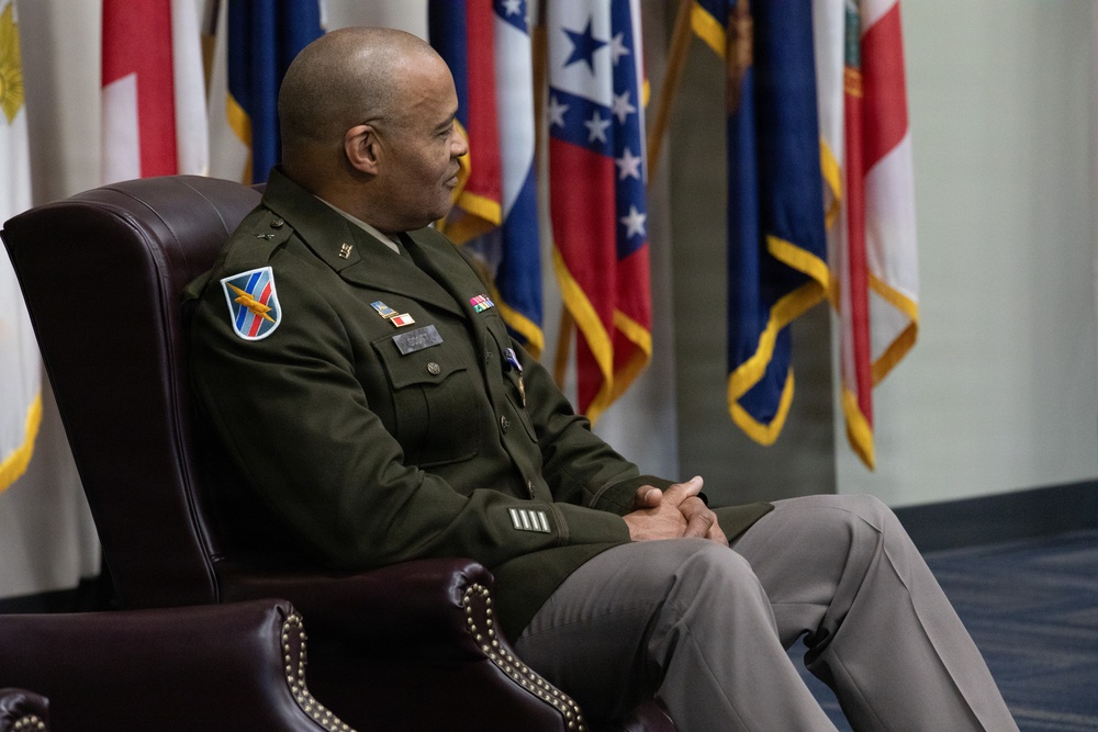 A U.S. Army officer in dress green uniform is seated in a leather chair in what appears to be an official military setting. Multiple flags are displayed in the background, including what appear to be U.S. state flags and possibly international flags. The officer appears to be listening or observing during a formal ceremony or official event.