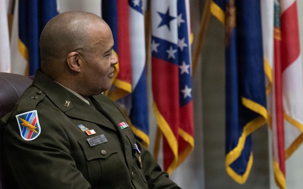 A U.S. Army officer in dress green uniform is seated in a leather chair in what appears to be an official military setting. Multiple flags are displayed in the background, including what appear to be U.S. state flags and possibly international flags. The officer appears to be listening or observing during a formal ceremony or official event.