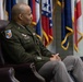 A U.S. Army officer in dress green uniform is seated in a leather chair in what appears to be an official military setting. Multiple flags are displayed in the background, including what appear to be U.S. state flags and possibly international flags. The officer appears to be listening or observing during a formal ceremony or official event.