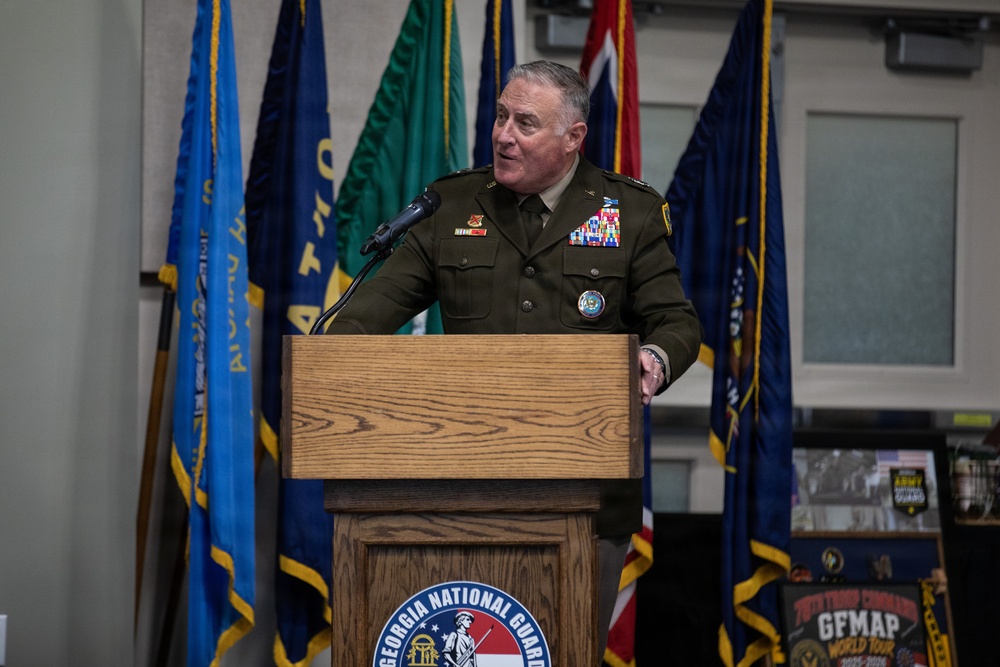 A senior military officer in U.S. Army dress greens uniform is speaking at a podium bearing the Georgia National Guard seal. The setting appears to be an official military ceremony or briefing, with multiple flags displayed in the background. The officer is wearing numerous medals, ribbons, and insignia indicating high rank and extensive service.