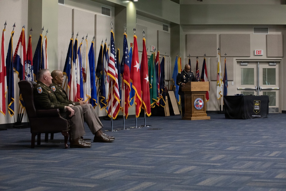A formal military ceremony is taking place in an indoor venue, likely an Army National Guard facility. A uniformed officer is speaking at a wooden podium bearing an Army National Guard seal, while two other uniformed officers are seated in chairs to the left. A large display of military and state flags forms a backdrop behind the seated officers.