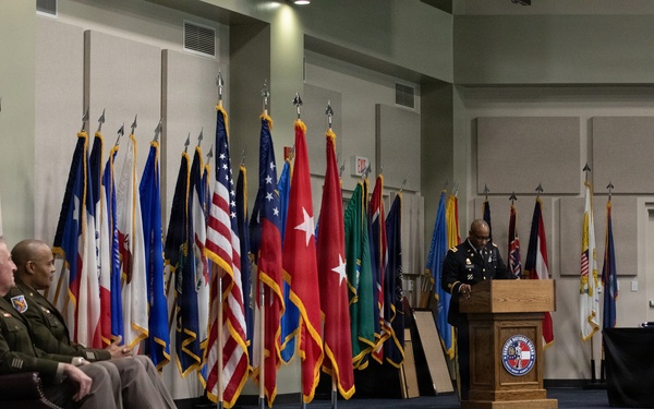A formal military ceremony is taking place in an indoor venue, likely an Army National Guard facility. A uniformed officer is speaking at a wooden podium bearing an Army National Guard seal, while two other uniformed officers are seated in chairs to the left. A large display of military and state flags forms a backdrop behind the seated officers.