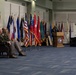 A formal military ceremony is taking place in an indoor venue, likely an Army National Guard facility. A uniformed officer is speaking at a wooden podium bearing an Army National Guard seal, while two other uniformed officers are seated in chairs to the left. A large display of military and state flags forms a backdrop behind the seated officers.