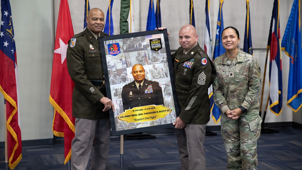 A military ceremony honoring U.S. Army Brigadier General Theodore R. Scott III, with three service members posing with a commemorative framed photo collage. The framed piece displays the dates '01 APR 1986 - 31 AUG 2026' and the motto 'Support the Fight,' along with the Georgia Army National Guard insignia. The ceremony appears to be a retirement or farewell event held in a formal military setting.