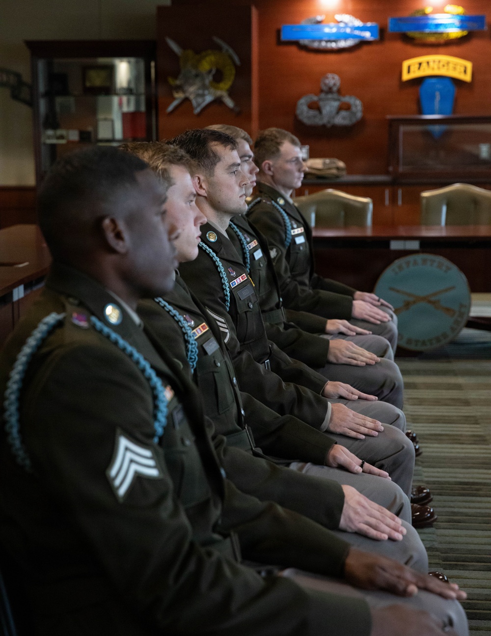 Five U.S. Army soldiers in dress green uniforms are seated in a row, appearing to be at attention during a formal ceremony or briefing. They are in a military office or conference room decorated with Army insignia, including a visible 'RANGER' tab, Infantry crossed rifles emblem, and Master Parachutist badge. The soldiers wear light blue shoulder cords (Infantry), service ribbons, and rank insignia.