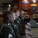 Five U.S. Army soldiers in dress green uniforms are seated in a row, appearing to be at attention during a formal ceremony or briefing. They are in a military office or conference room decorated with Army insignia, including a visible 'RANGER' tab, Infantry crossed rifles emblem, and Master Parachutist badge. The soldiers wear light blue shoulder cords (Infantry), service ribbons, and rank insignia.
