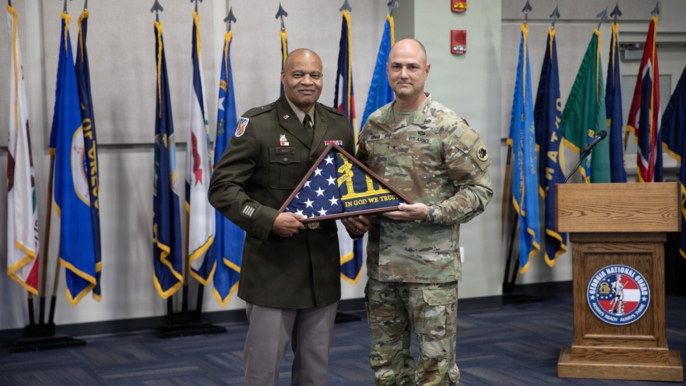 Two U.S. Army personnel are posing together at a formal military ceremony, jointly holding a framed folded American flag with an inscription reading 'In God We Trust.' The event appears to be a retirement, award, or recognition ceremony taking place at a Georgia National Guard facility, as indicated by the podium seal visible on the right. Multiple U.S. state and military branch flags are displayed in the background.