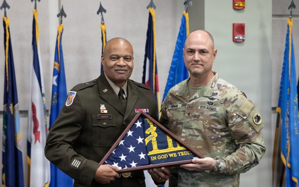 Two U.S. Army personnel are posing together at a formal military ceremony, jointly holding a framed folded American flag with an inscription reading 'In God We Trust.' The event appears to be a retirement, award, or recognition ceremony taking place at a Georgia National Guard facility, as indicated by the podium seal visible on the right. Multiple U.S. state and military branch flags are displayed in the background.