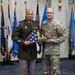 Two U.S. Army personnel are posing together at a formal military ceremony, jointly holding a framed folded American flag with an inscription reading 'In God We Trust.' The event appears to be a retirement, award, or recognition ceremony taking place at a Georgia National Guard facility, as indicated by the podium seal visible on the right. Multiple U.S. state and military branch flags are displayed in the background.