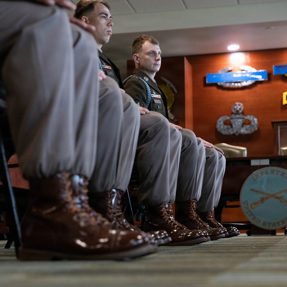 Several military personnel in dress uniforms are seated in a formal indoor setting, likely during a briefing or ceremony. They wear olive/tan Army Service Uniforms with highly polished brown dress boots. The background displays military insignia, badges, and a partially visible 'Infantry, United States' seal on the wall.