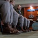 Several military personnel in dress uniforms are seated in a formal indoor setting, likely during a briefing or ceremony. They wear olive/tan Army Service Uniforms with highly polished brown dress boots. The background displays military insignia, badges, and a partially visible 'Infantry, United States' seal on the wall.