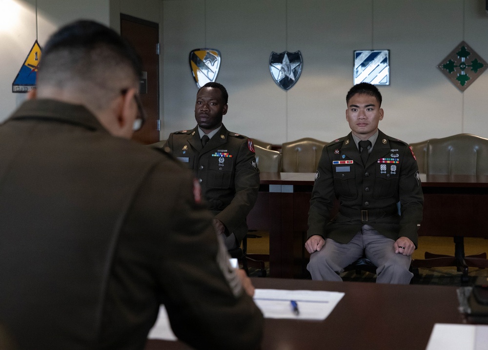 Three U.S. Army soldiers in Army Service Uniforms (ASUs) are gathered in a formal military conference or board room. Two soldiers are seated facing a third soldier whose back is to the camera, suggesting a formal board or evaluation proceeding. Military division insignia plaques are mounted on the wall behind them.