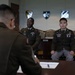 Three U.S. Army soldiers in Army Service Uniforms (ASUs) are gathered in a formal military conference or board room. Two soldiers are seated facing a third soldier whose back is to the camera, suggesting a formal board or evaluation proceeding. Military division insignia plaques are mounted on the wall behind them.
