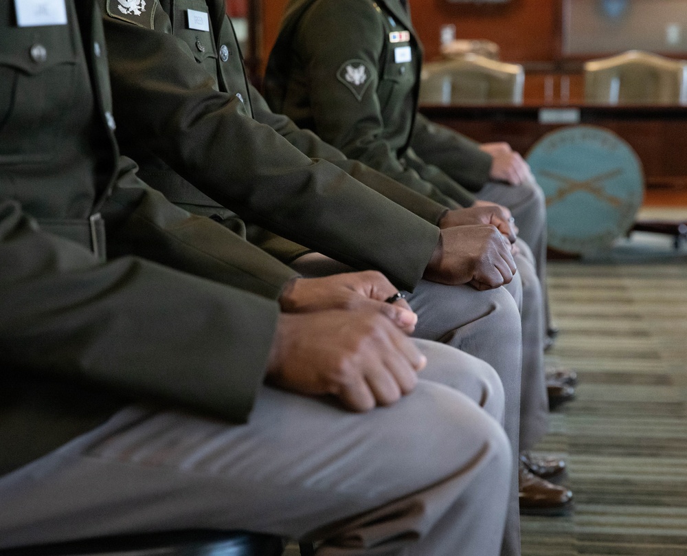 The image shows a row of military personnel seated in formal dress uniforms, viewed from a side angle focusing on their torsos, laps, and hands. The individuals are wearing olive/dark green Army service uniforms with visible insignia, patches, and name tags. The setting appears to be an official indoor military venue with formal furniture visible in the background.