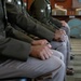 The image shows a row of military personnel seated in formal dress uniforms, viewed from a side angle focusing on their torsos, laps, and hands. The individuals are wearing olive/dark green Army service uniforms with visible insignia, patches, and name tags. The setting appears to be an official indoor military venue with formal furniture visible in the background.