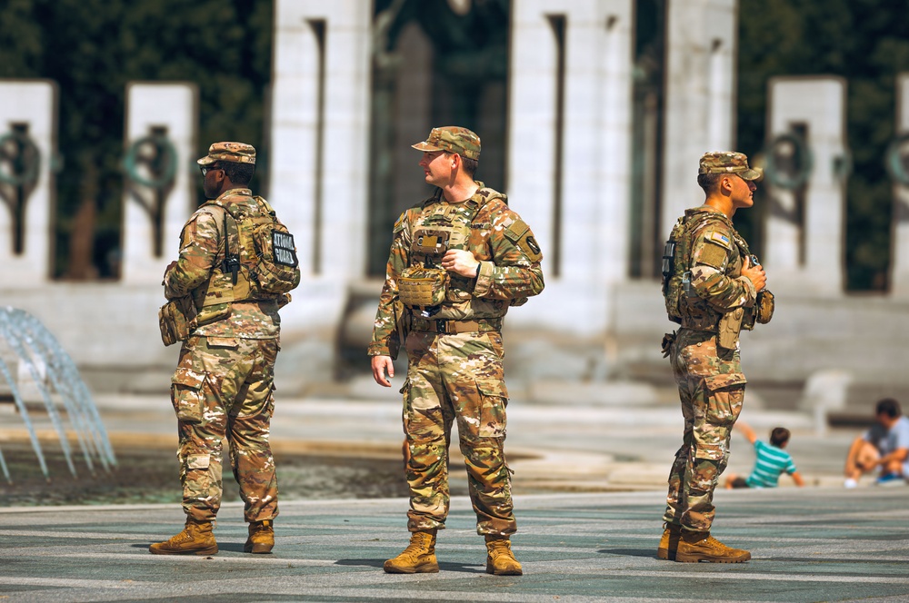 Three soldiers in U.S. Army MultiCam (OCP) camouflage uniforms are standing at what appears to be the National World War II Memorial in Washington, D.C. The patch on the back of the leftmost soldier's vest reads 'NATIONAL GUARD,' identifying the unit. Civilians, including children, are visible in the background relaxing near the memorial.