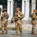 Three soldiers in U.S. Army MultiCam (OCP) camouflage uniforms are standing at what appears to be the National World War II Memorial in Washington, D.C. The patch on the back of the leftmost soldier's vest reads 'NATIONAL GUARD,' identifying the unit. Civilians, including children, are visible in the background relaxing near the memorial.